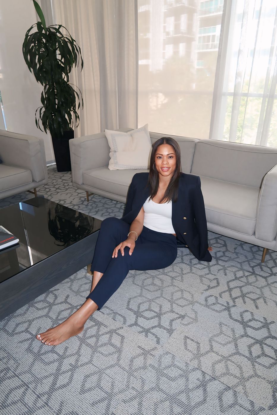 Cara Coley seated on the floor of a sunlit lounge in a navy suit